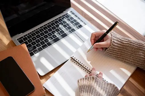 a-top-view-image-of-a-woman-is-taking-notes-in-her-notebook-while-working-on-her-laptop