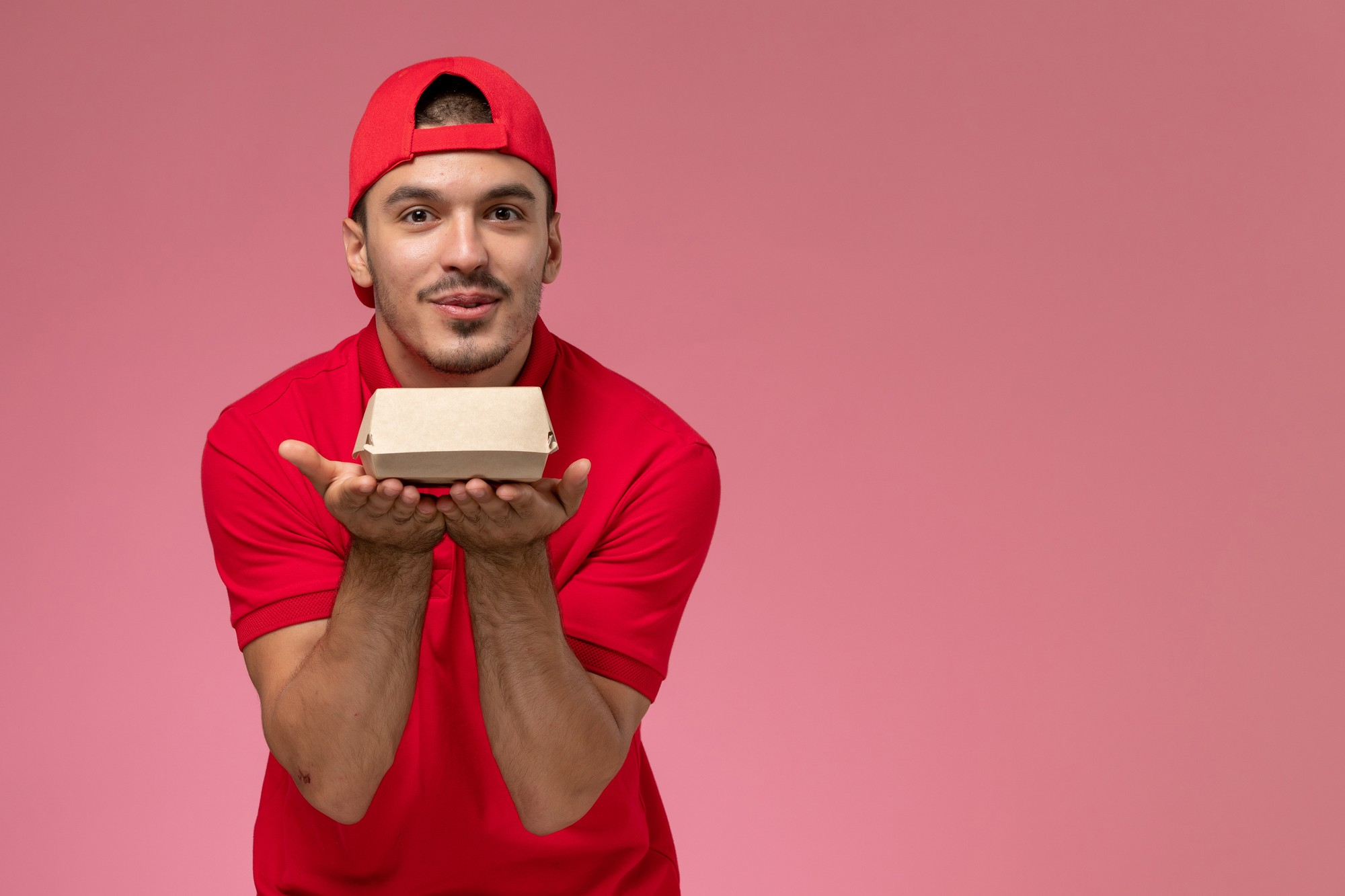 front-view-male-courier-red-uniform-cape-holding-little-delivery-package-pink-background