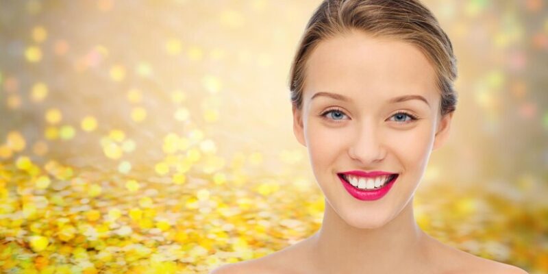 beauty-people-health-concept-smiling-young-woman-face-with-pink-lipstick-lips-shoulders-golden-glitter-holidays-lights-background_380164-113766